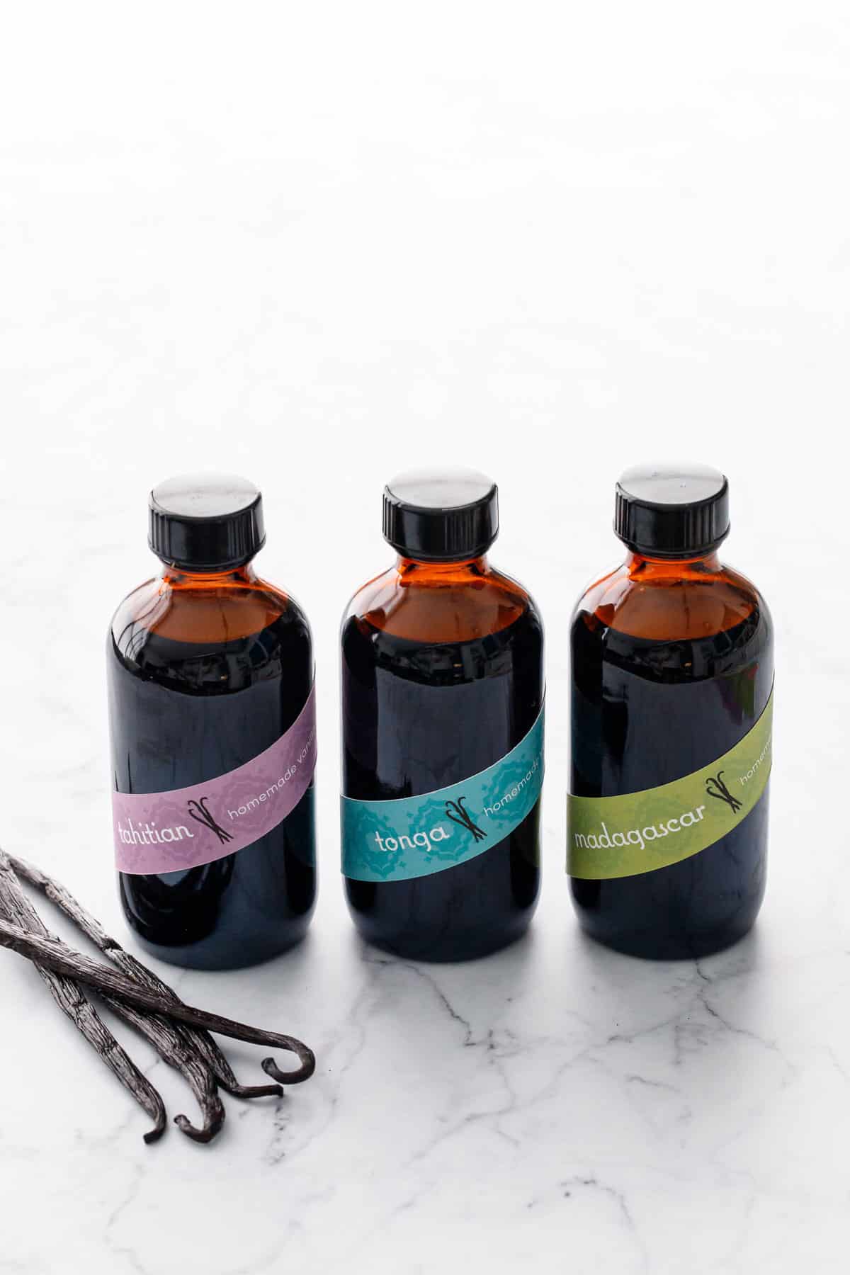 Three amber glass bottles filled with Homemade Vanilla Extract and colorful labels, with a few vanilla beans on the side.