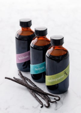 Row of three amber glass bottles filled with Homemade Vanilla Extract, with colorful diagonal band labels and a few vanilla beans on the side.