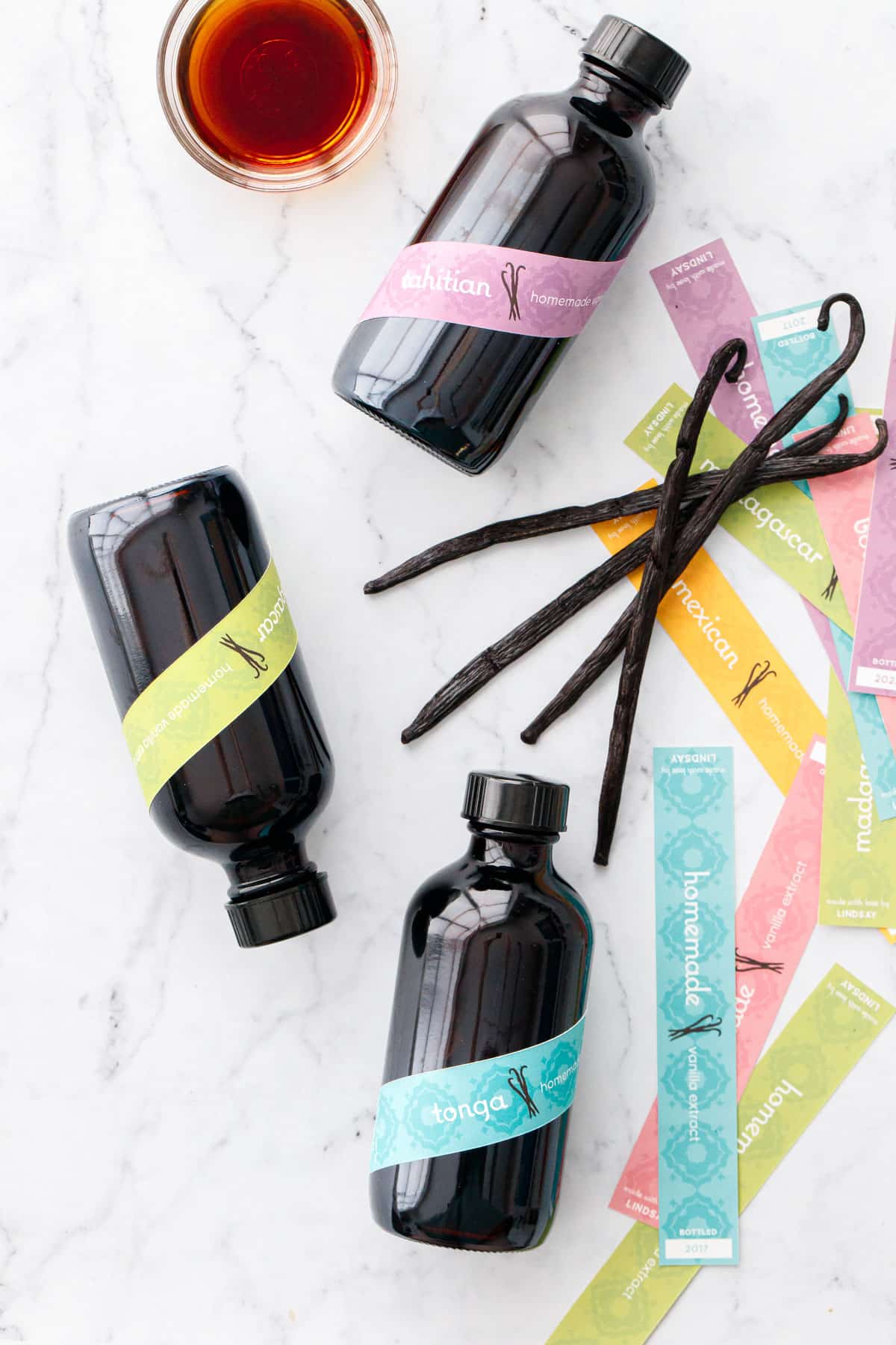 Overhead, three bottles of Homemade Vanilla Extract with colorful band labels, with a small bowl of vanilla extract and a few fresh vanilla bean pods in a pile.