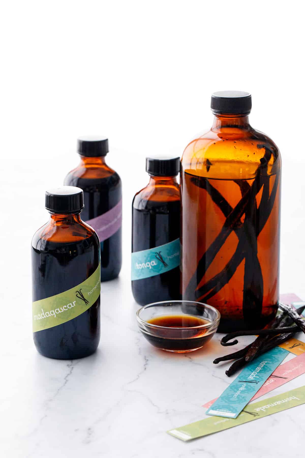 Bottles of freshly made Homemade Vanilla Extract with colorful labels, and a bottle of alcohol backlit so you can see the beans inside, plus a small bowl of extract and a few beans on the side.
