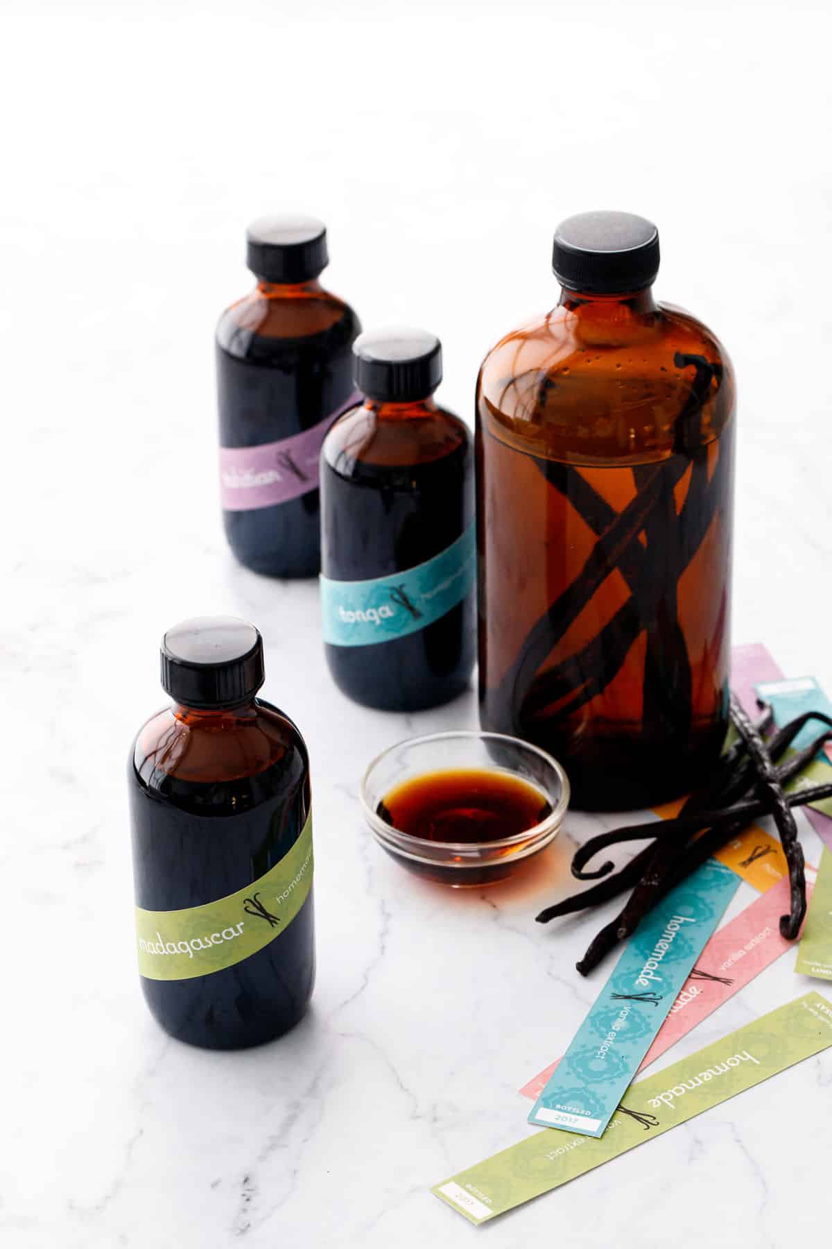 Bottles of Homemade Vanilla Extract in various stages of the process, one large bottle showing the before, and three small bottles with labels plus a small bowl of extract showing the final product.