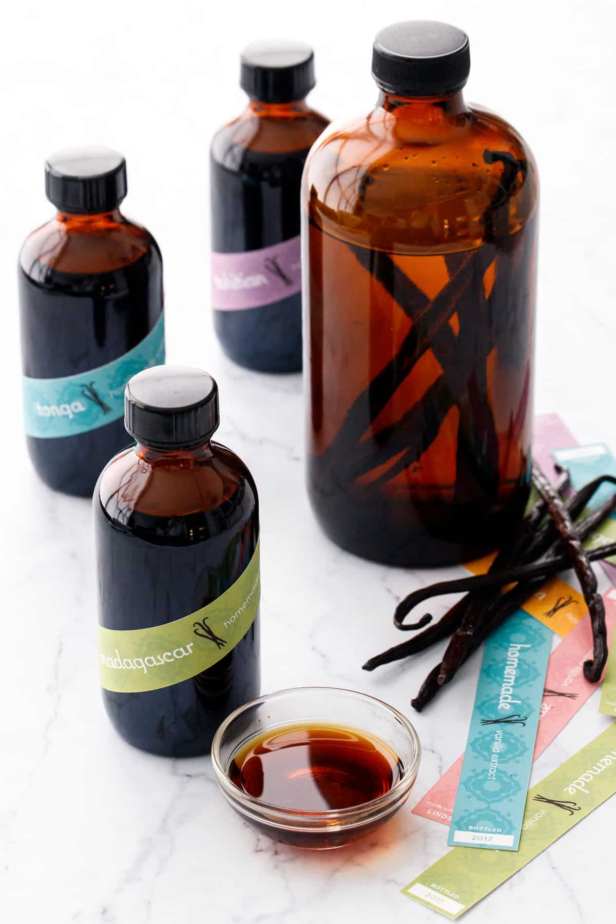 Homemade Vanilla Extract in amber glass bottles, three small ones with colorful labels and one large bottle backlit to show the beans submerged in alcohol before infusing.