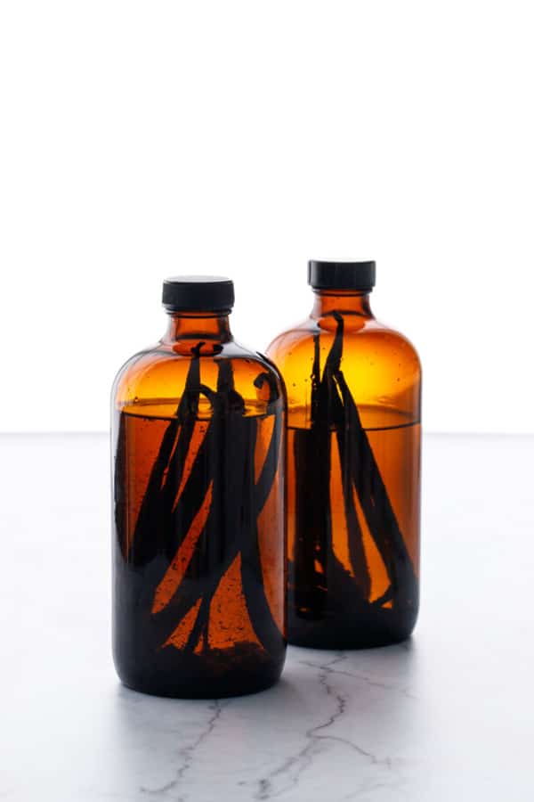 Backlit photo showing to amber glass bottles filled with alcohol and multiple vanilla beans submerged inside.