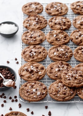 Rows of Nutella Chocolate Chip Cookies on a wire baking rack, with bowls of chocolate chips, flake salt, and a spoonful of Nutella on the side.