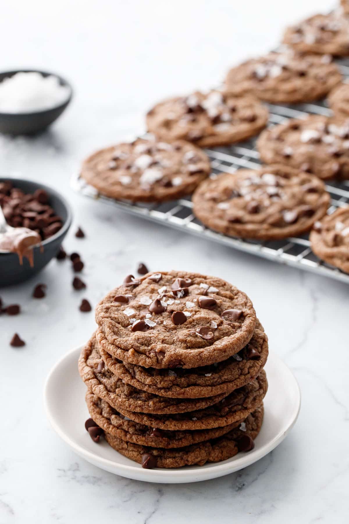Stack of Nutella Chocolate Chip Cookies on a small white plate, with a cooling rack with more cookies plus a bowl of chocolate chips and sea salt in the background.