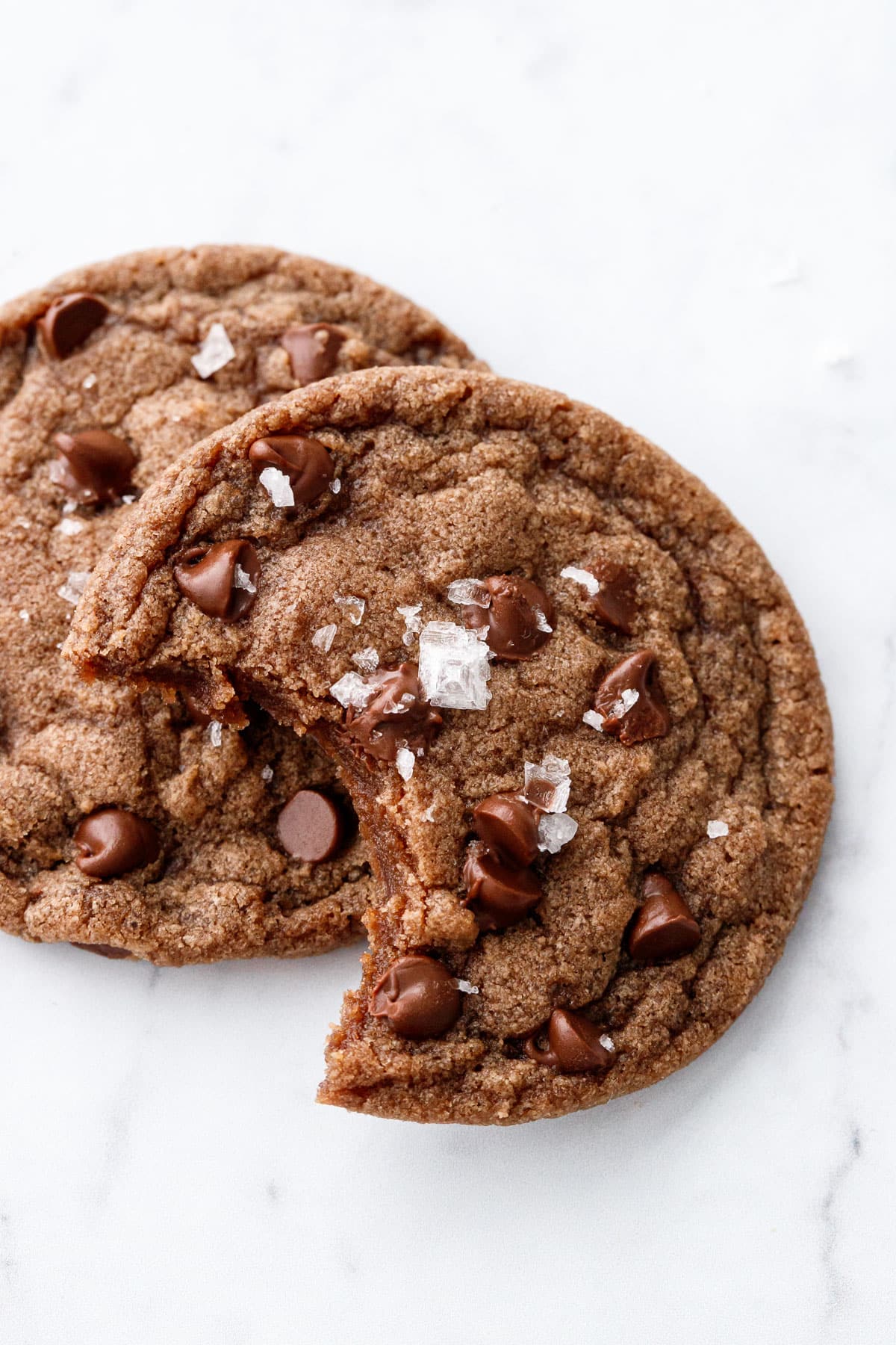 Two Nutella Chocolate Chip Cookies, one with a bite out of it, viewed close up to show the texture and flakes of sea salt on top.