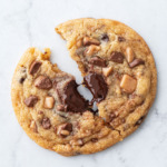 Closeup of a single large Toffee Chocolate Chip Cookie split in half, strand of melted chocolate pulling between the halves.