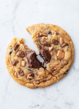 Closeup of a single large Toffee Chocolate Chip Cookie split in half, strand of melted chocolate pulling between the halves.