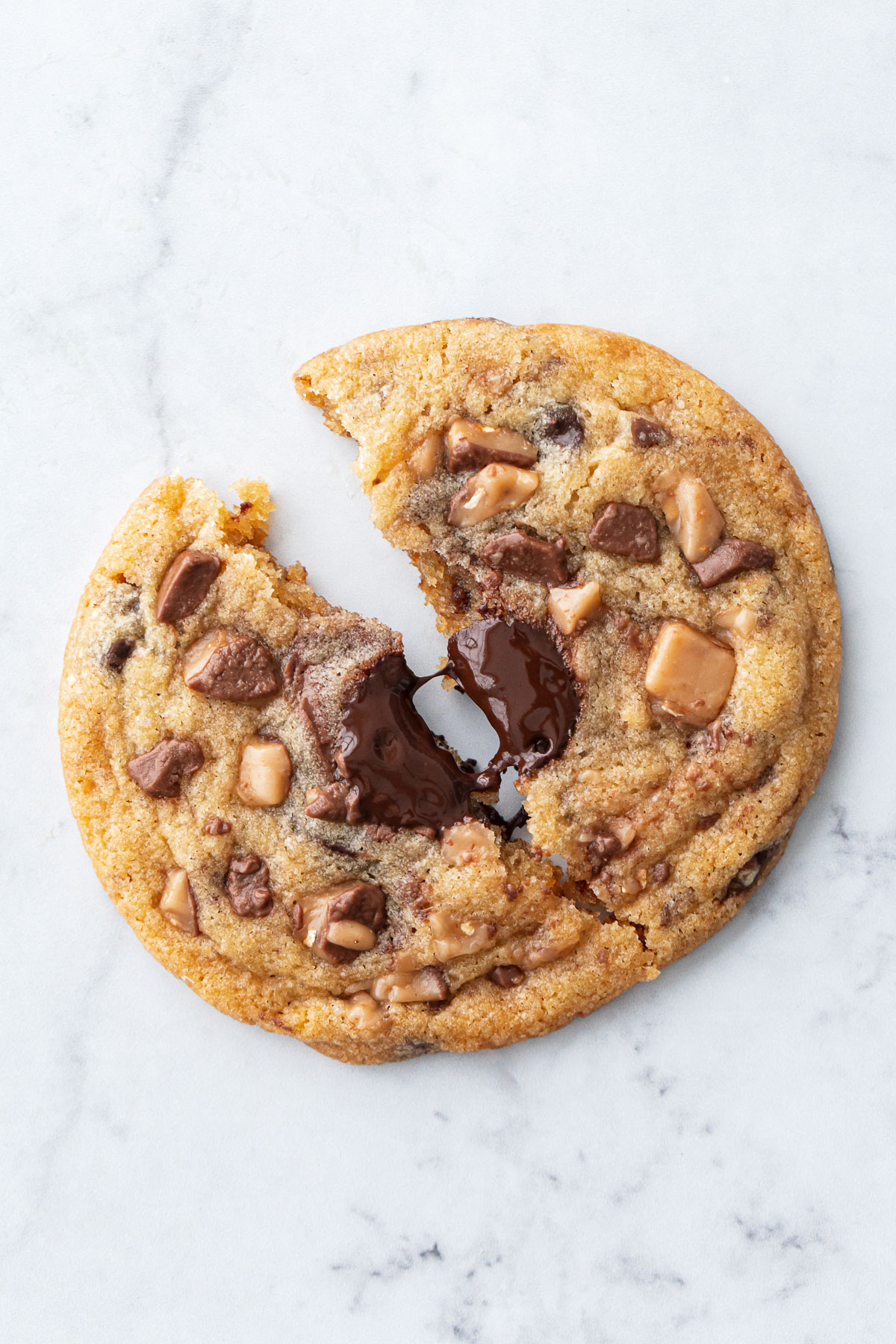 Closeup of a single large Toffee Chocolate Chip Cookie split in half, strand of melted chocolate pulling between the halves.