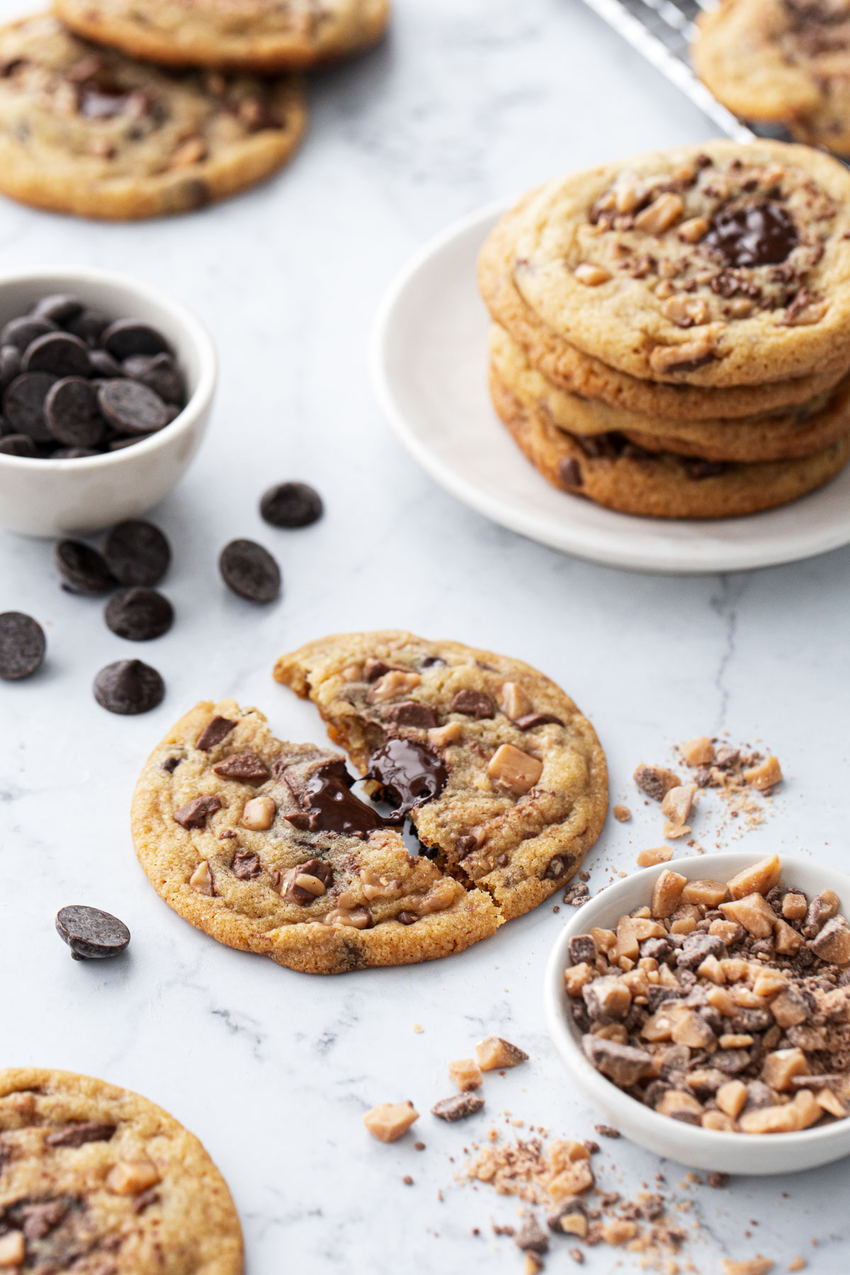 Toffee Chocolate Chip Cookies scattered on a marble surface with a bowl of toffee bits and bowl of chocolate chips in the background.