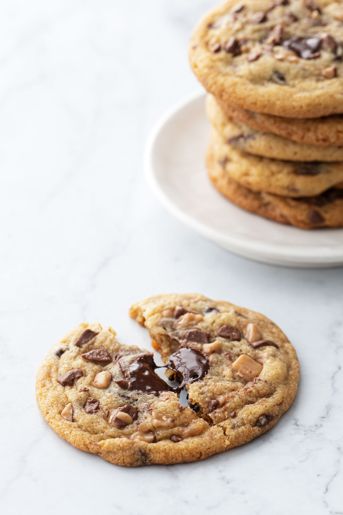 Toffee Chocolate Chip Cookie split in half to show the gooey interior texture, stack of cookies in the background.