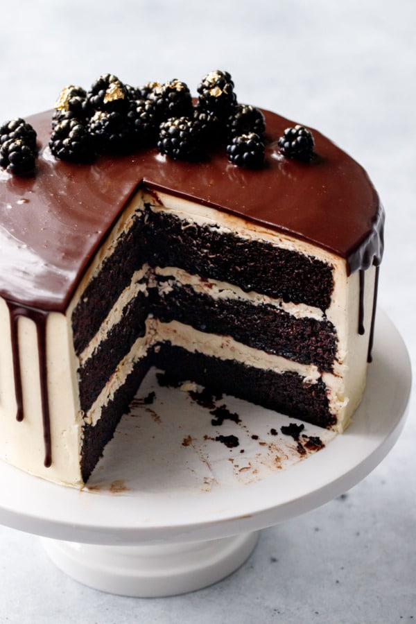 Blackberry Red Wine Chocolate Cake on a white cake stand with a wedge cut out of it to show the distinct layers inside.