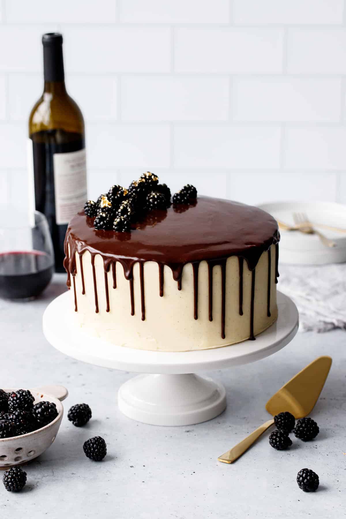 Blackberry Red Wine Chocolate Cake on a white cake plate, with glass and bottle of red wine and fresh blackberries in the backround.