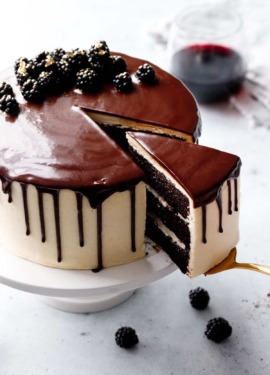 Gold cake server pulling out a slice of Blackberry Red Wine Chocolate Cake, with a shiny chocolate drippy glaze and decorated with gold-dusted blackberries on top.