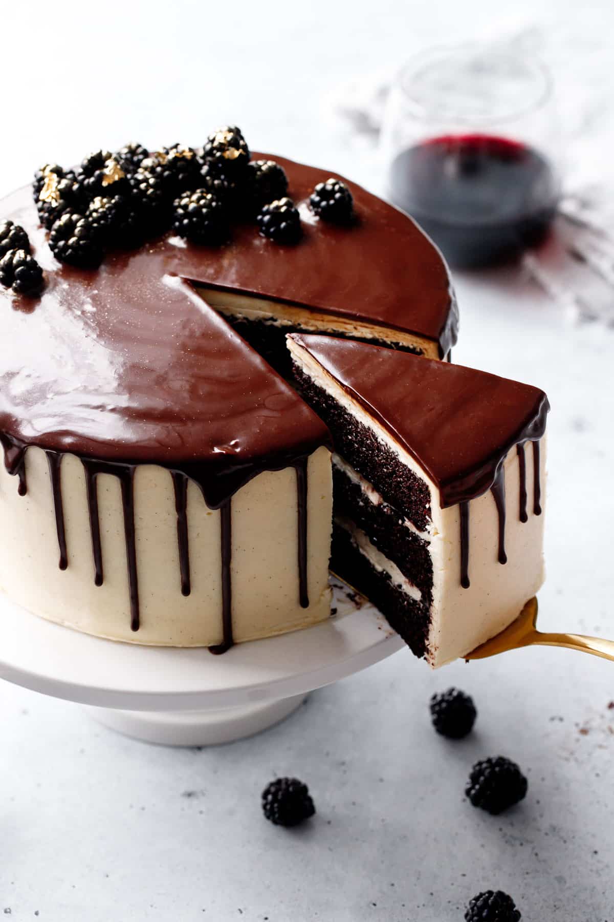 Gold cake server pulling out a slice of Blackberry Red Wine Chocolate Cake, with a shiny chocolate drippy glaze and decorated with gold-dusted blackberries on top.