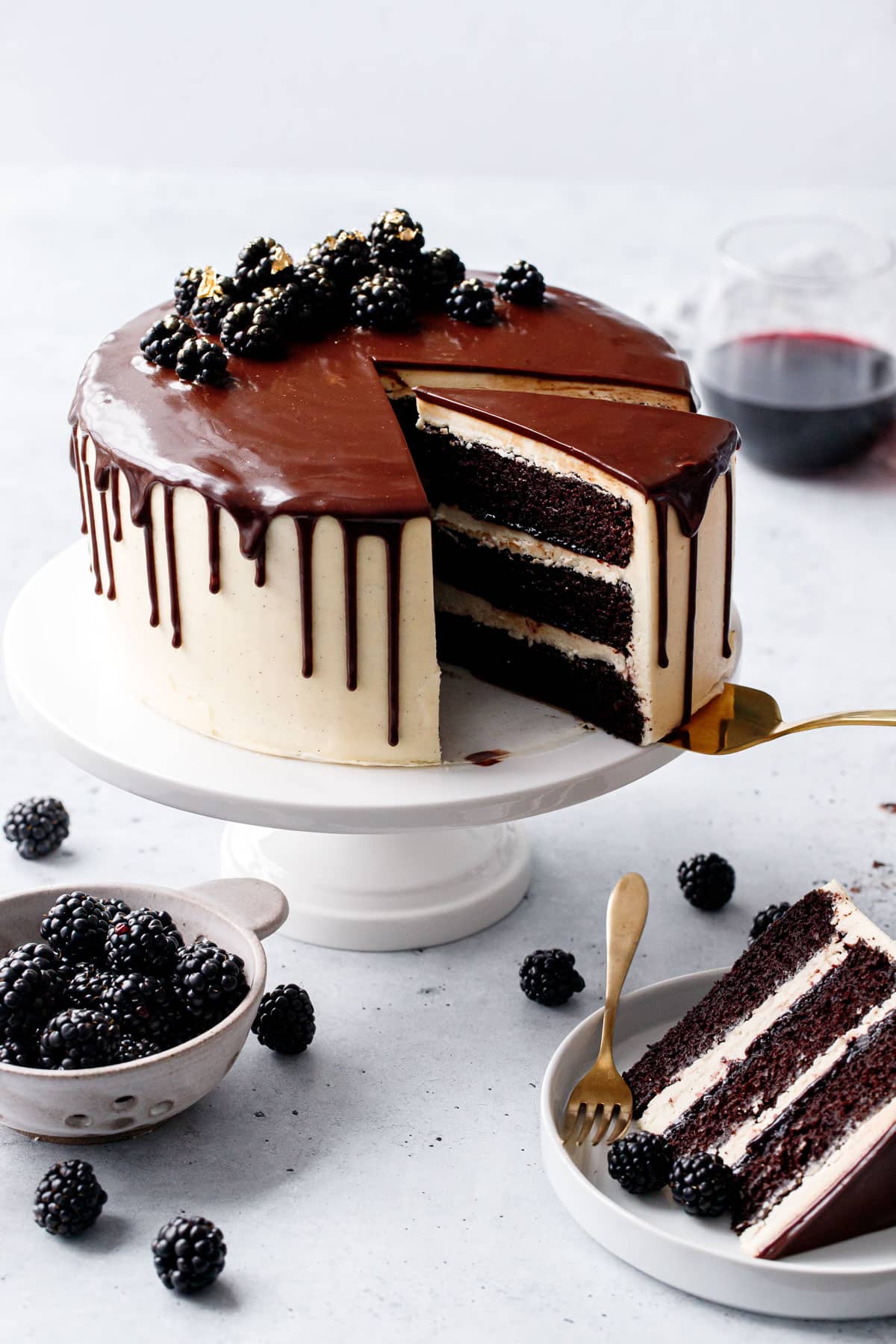 Sliced Blackberry Red Wine Chocolate Cake on a white cake stand, one slice on a plate to the side with a bowl of fresh blackberries, glass of red wine in the background.