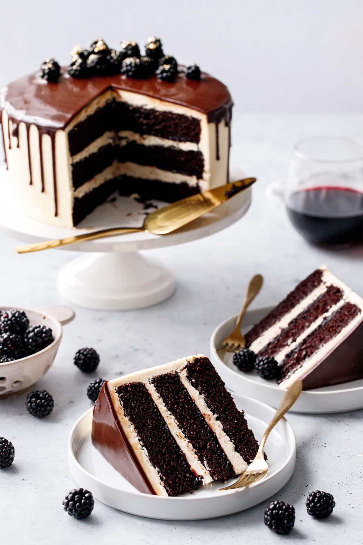 Slices of Blackberry Red Wine Chocolate Cake on white plates, whole cake with a gold cake server, glass of red wine, and fresh blackberries in the background.