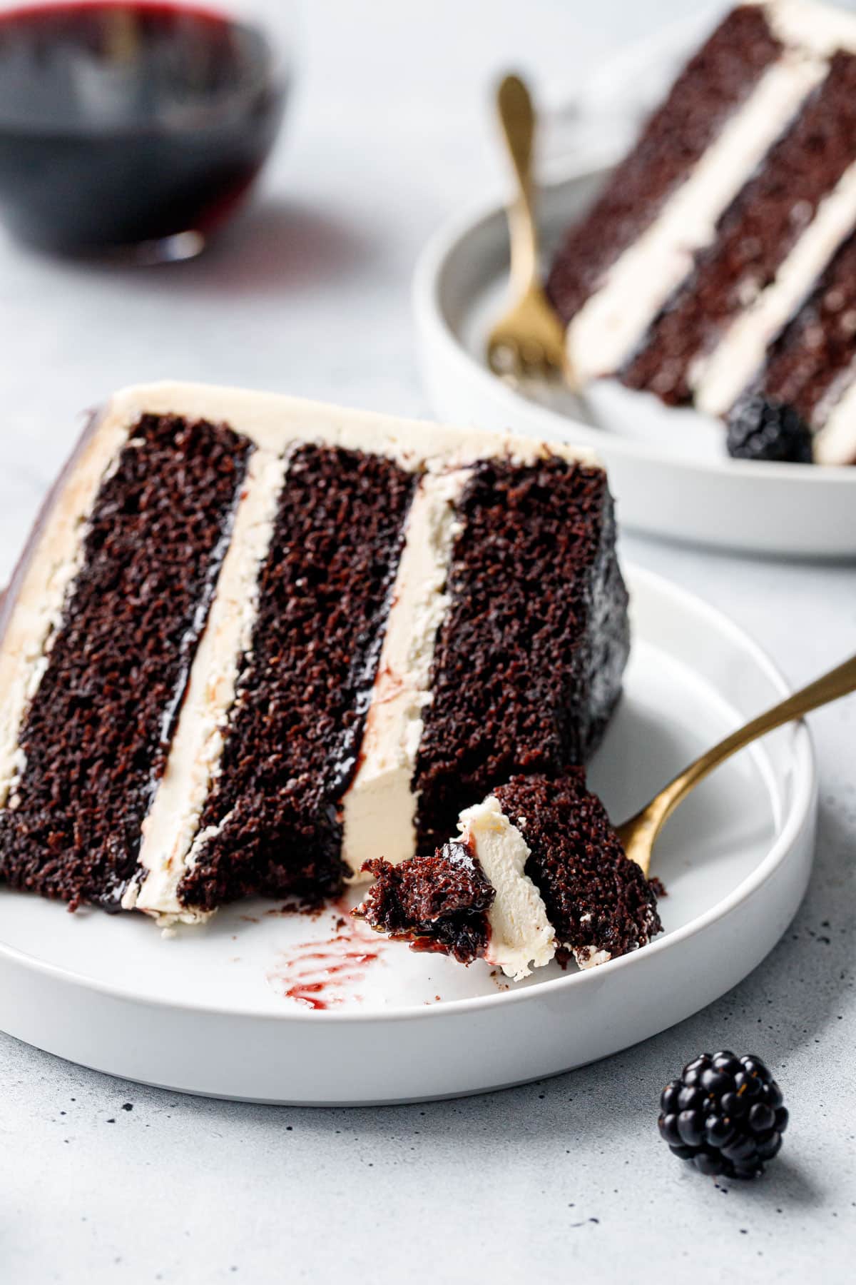 Closeup, slice of Blackberry Red Wine Chocolate Cake with a forkful taken out of it, showing the distinct layers, moist crumb, and blackberry jam hidden in between layers.