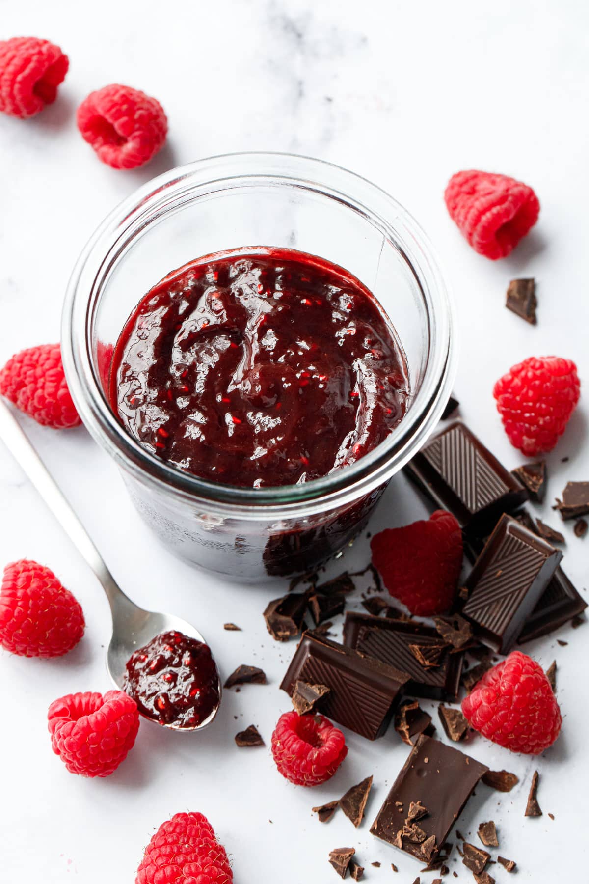 Jar of Chocolate Raspberry Jam with chopped squares of chocolate and fresh raspberries scattered around, with a spoonful of jam on the side.