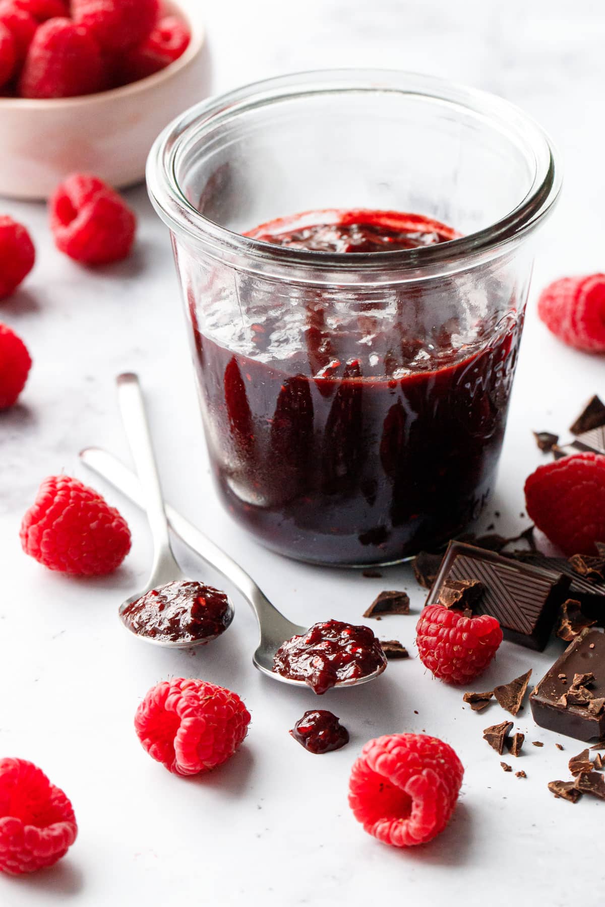 Glass jar filled with Chocolate Raspberry Jam, two spoons of jam plus chopped chocolate and fresh raspberries scattered around on a marble background.