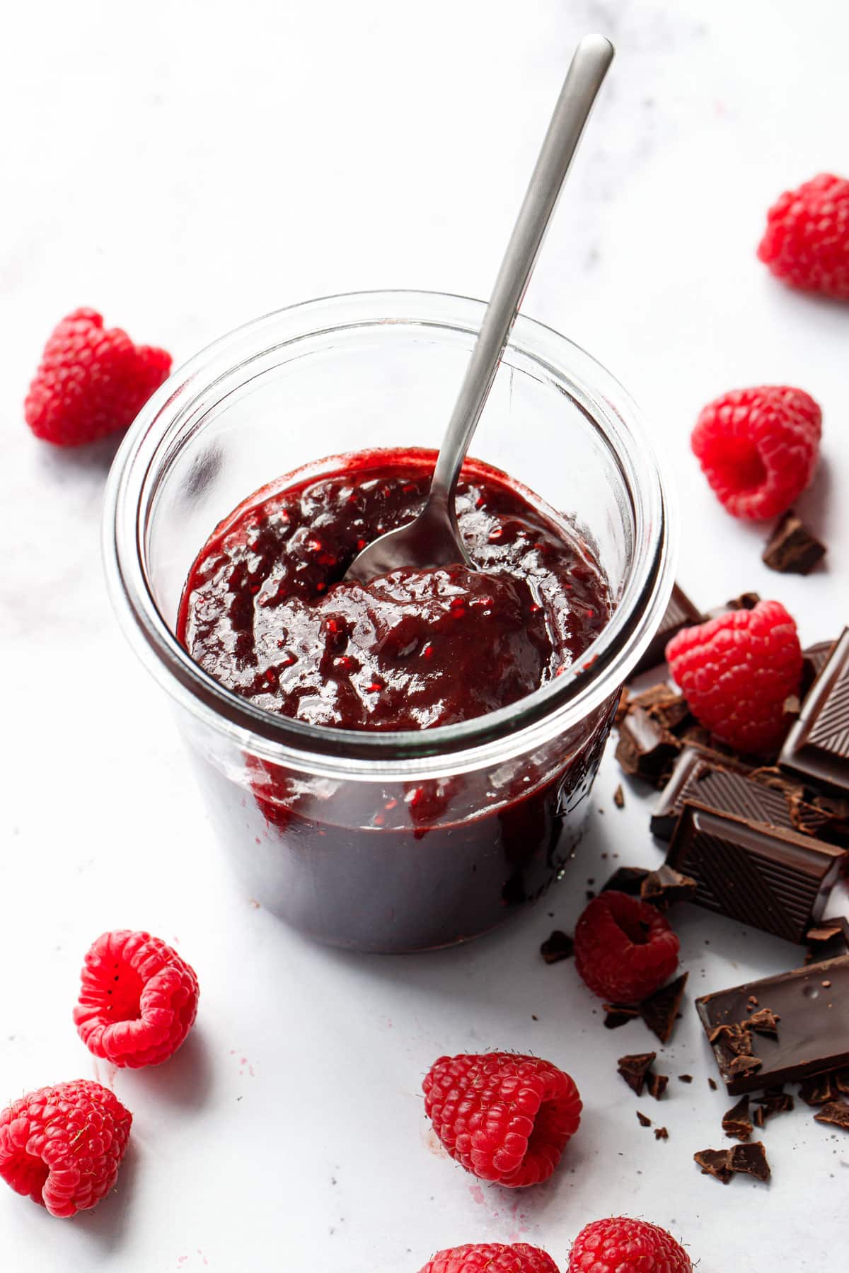 Swirl of Chocolate Raspberry Jam in a glass jar with silver spoon, fresh raspberries and squares of dark chocolate in the background.