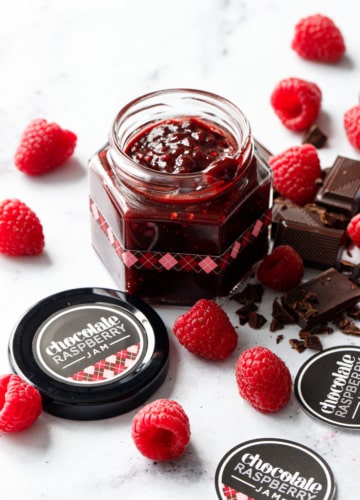 Small faceted glass canning jar filled with Chocolate Raspberry Jam, with brown and pink argyle designed labels and chopped chocolate and fresh raspberries scattered around.