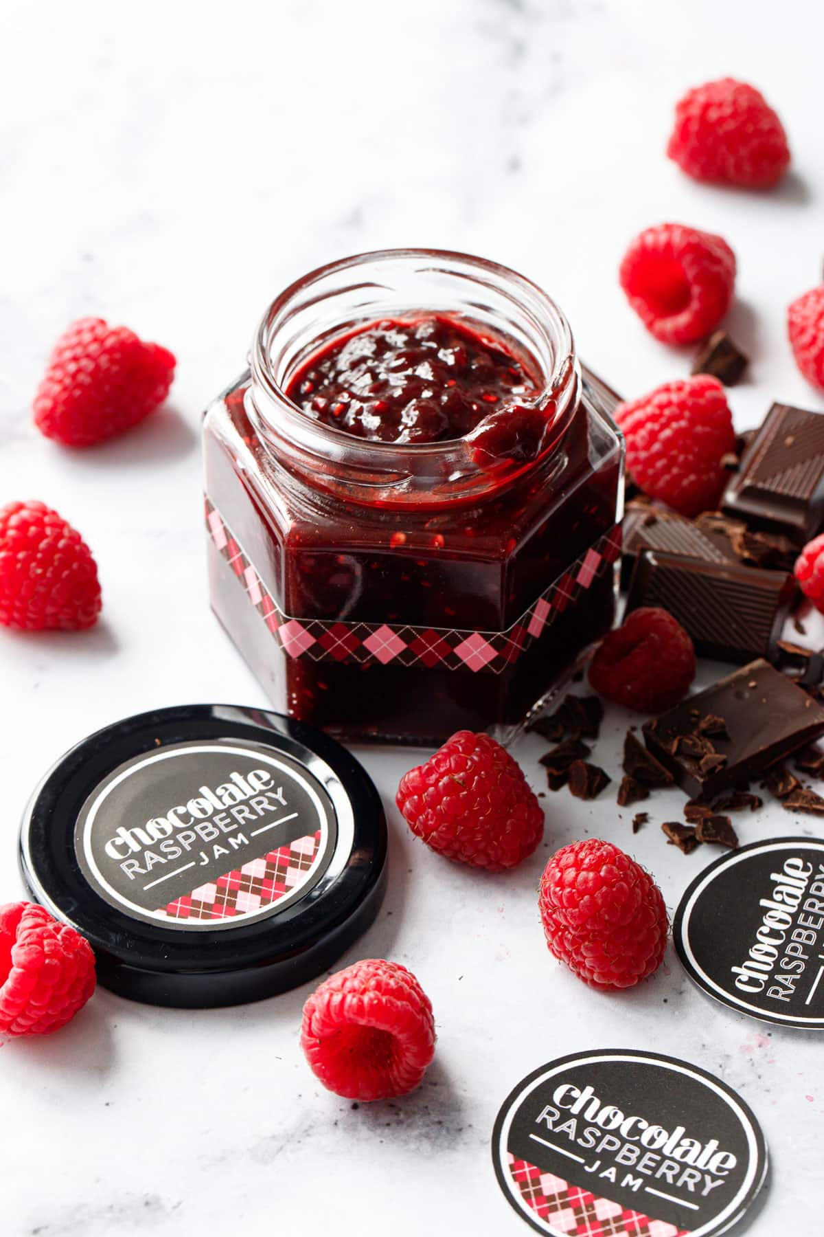 Small faceted glass canning jar filled with Chocolate Raspberry Jam, with brown and pink argyle designed labels and chopped chocolate and fresh raspberries scattered around.
