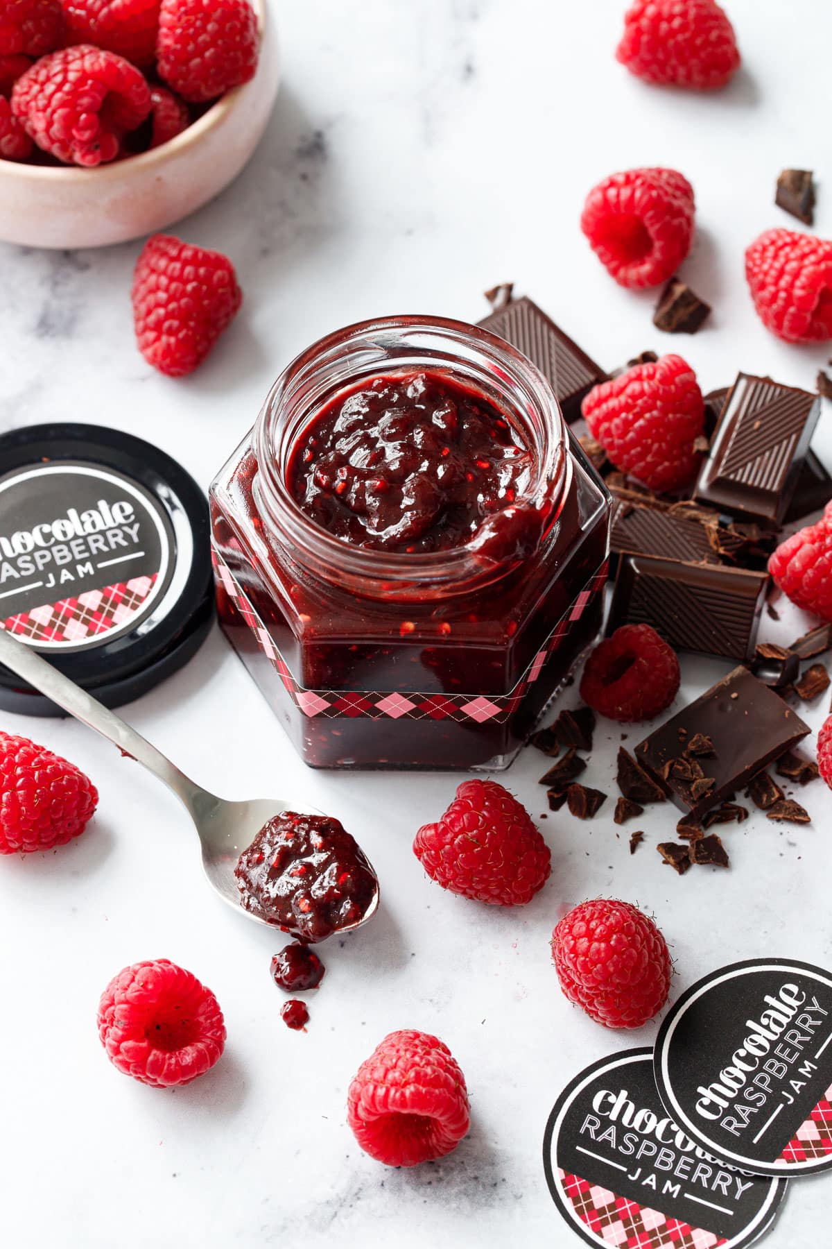 Chocolate Raspberry Jam in a hexagon glass jar, black lids with brown and pink argyle designed labels and chopped chocolate and fresh raspberries scattered around.