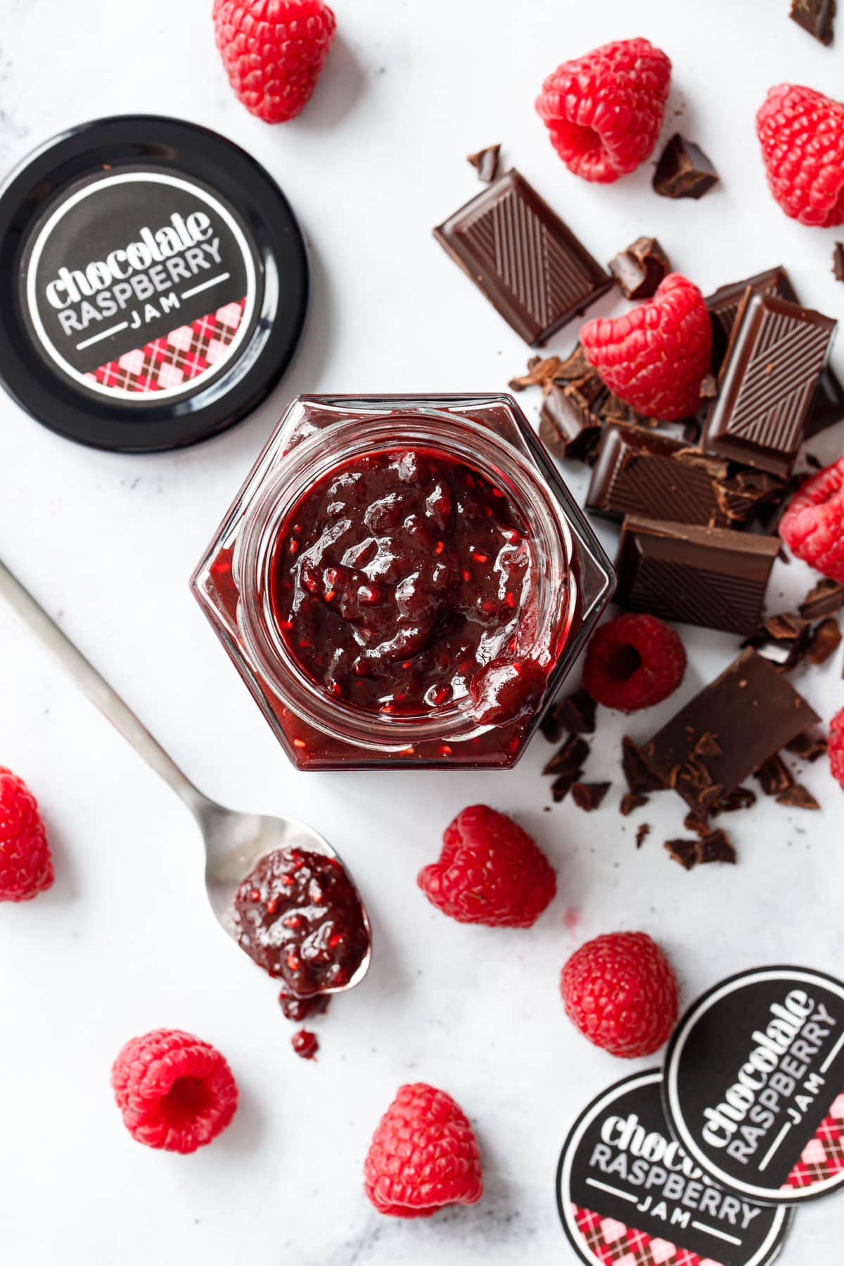 Overhead, jar filled with Chocolate Raspberry Jam, with spoonful of jam, printed jam labels, chopped chocolate and fresh raspberries messily arranged on a marble background.