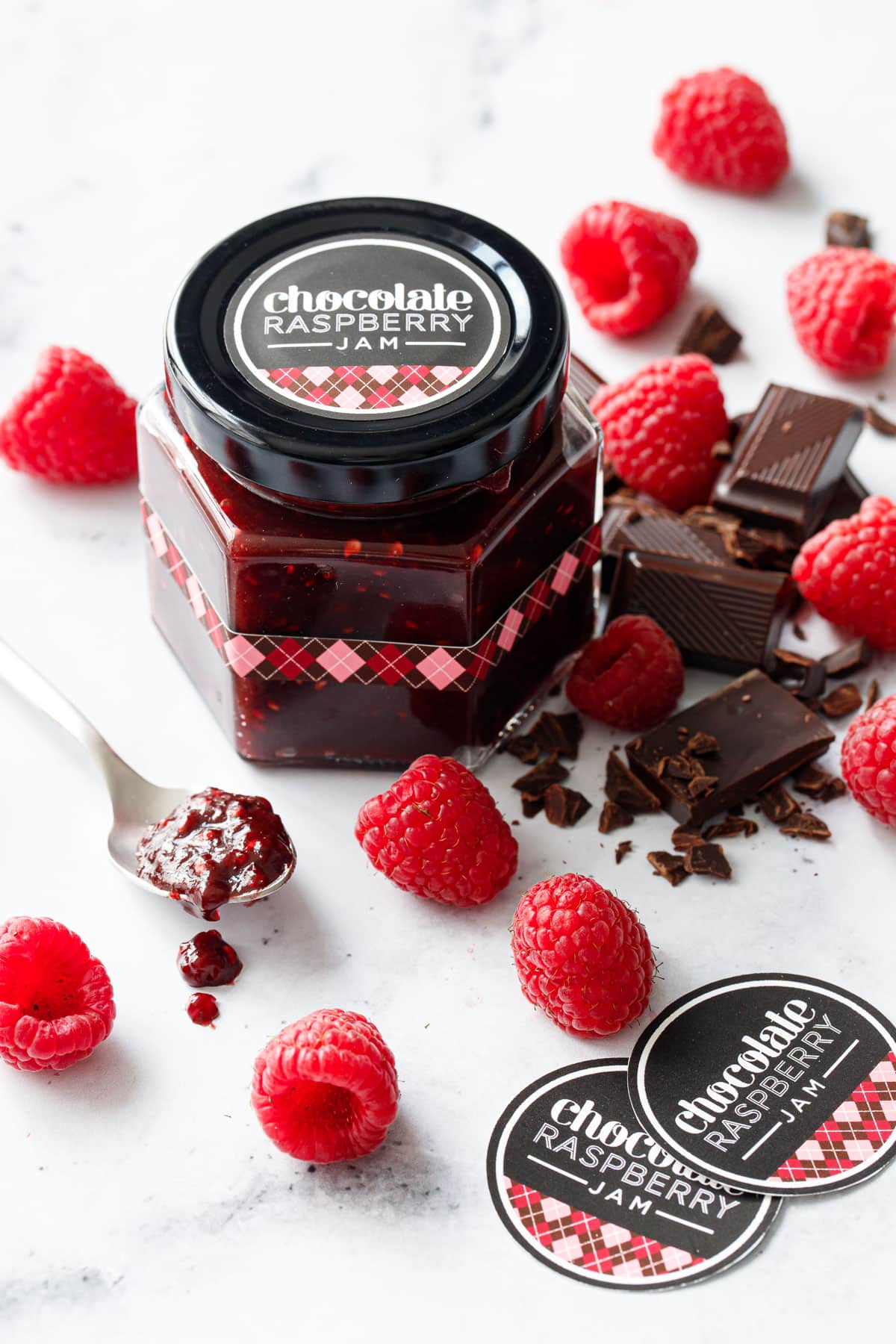 Jar of Chocolate Raspberry Jam with pink and brown printed label design, plus a spoon and fresh raspberries and dark chocolate.