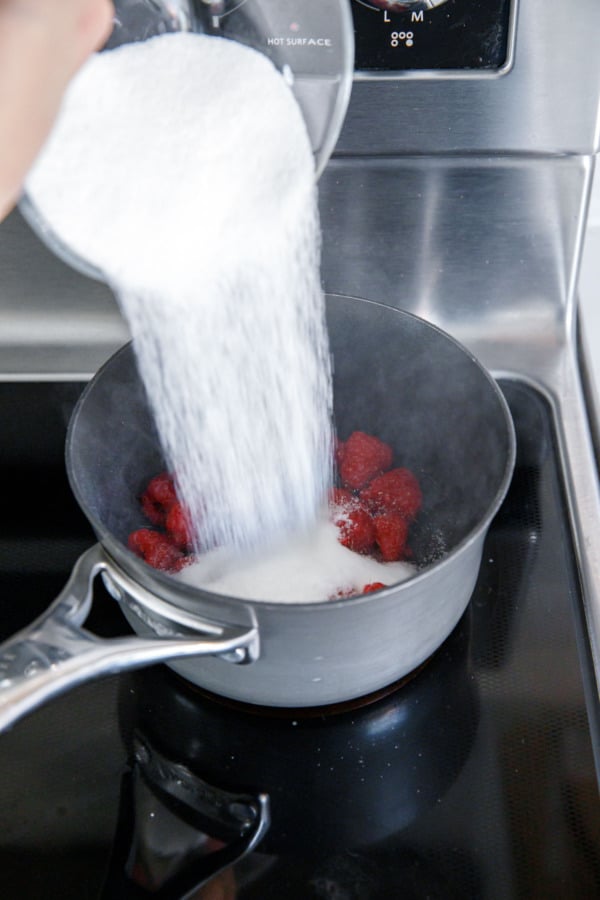 Pouring sugar into a pot of raspberries on the stovetop.