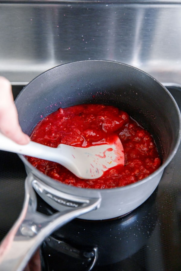 Mashing raspberries with a spatula while cooking them in a small saucepan on an electric stovetop.
