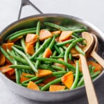 Stainless skillet with Sautéed Green Beans and Persimmons and wooden serving utensils on a gray background.