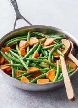 Stainless skillet with Sautéed Green Beans and Persimmons and wooden serving utensils on a gray background.