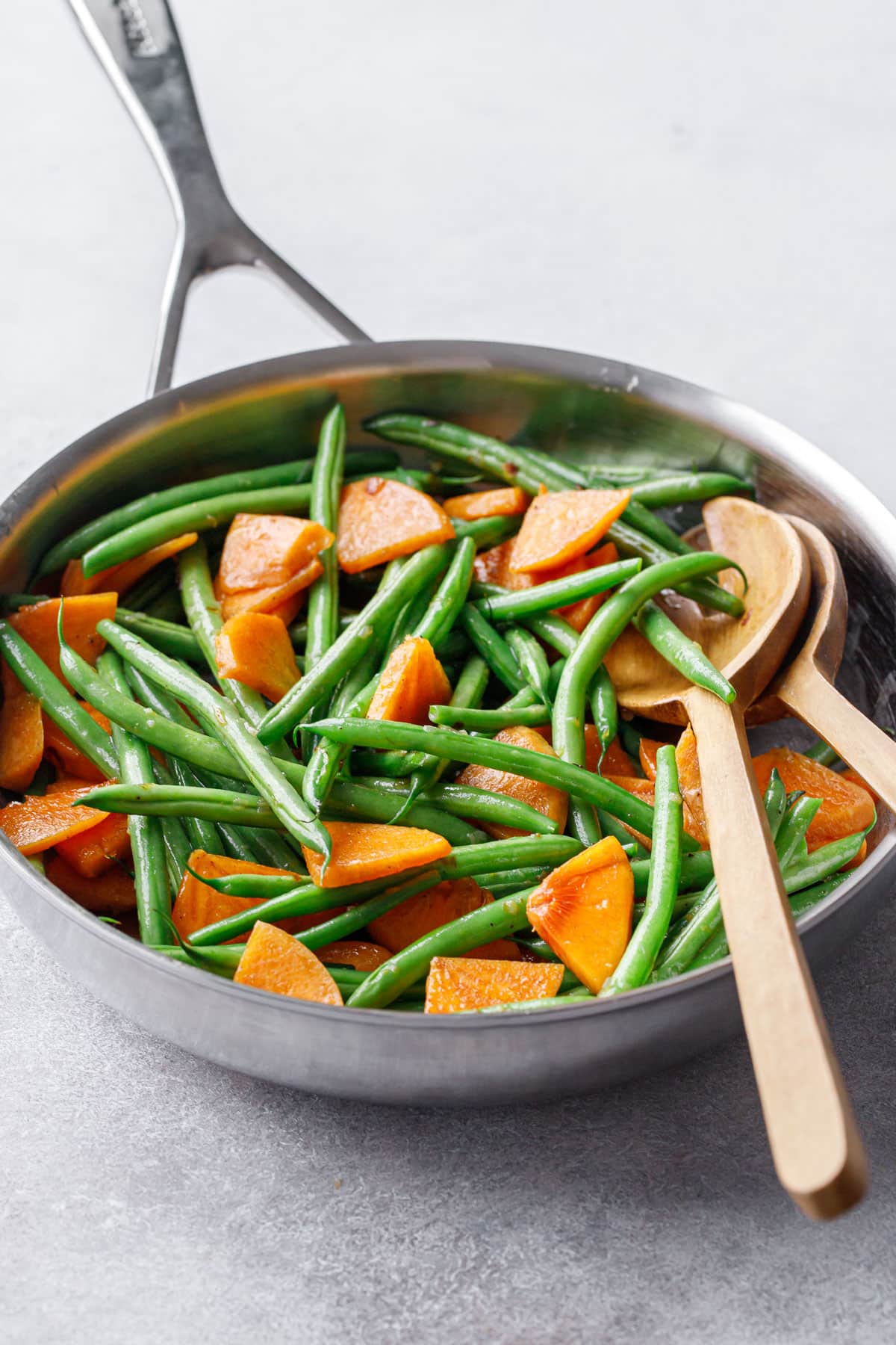 Stainless skillet with Sautéed Green Beans and Persimmons and wooden serving utensils on a gray background.