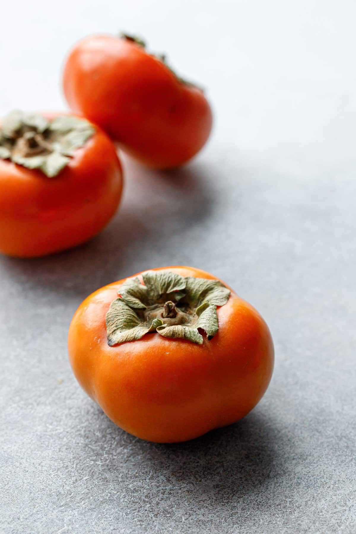 Three Fuyu persimmons that look a lot like orange tomatoes on a gray background.