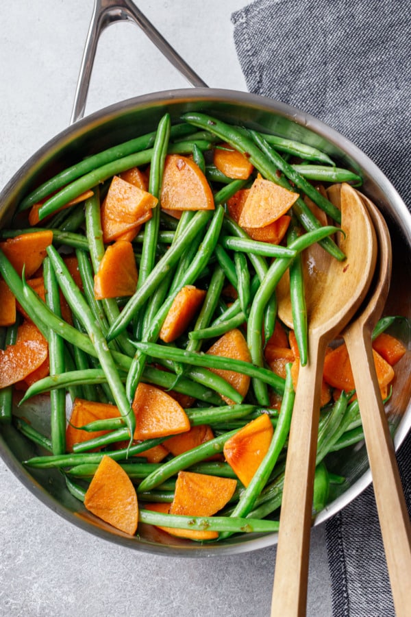 Overhead view, stainless skillet with Sautéed Green Beans and Persimmons, with wooden serving utensils and a gray denim napkin.