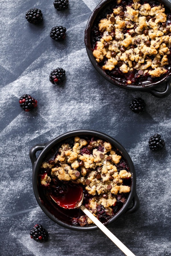 Overhead, two black baking dishes with Blackberry Chocolate Chip Cookie Crumble, one with a spoonful to show juicy berry filling