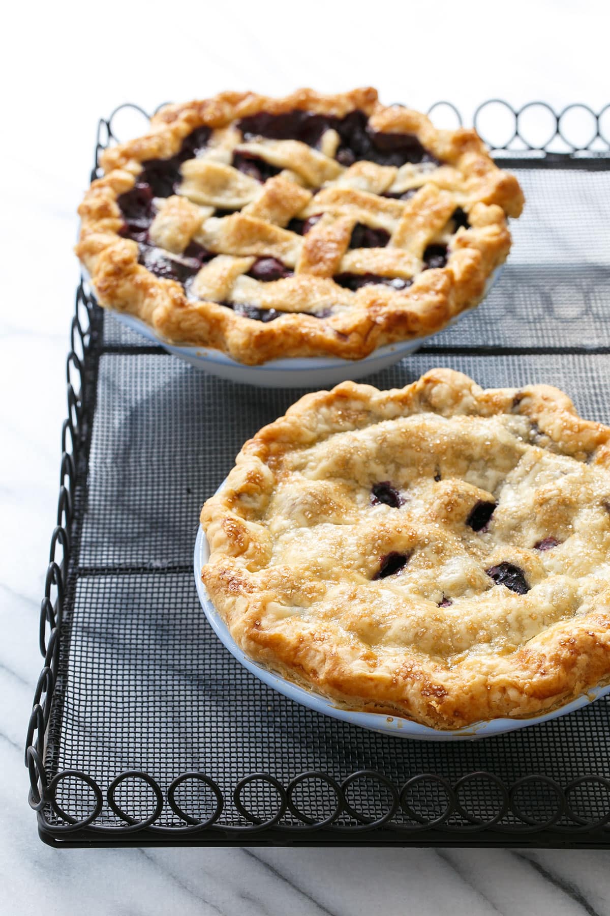 Two mini fresh blueberry pies on a wire rack, backlit with marble background.