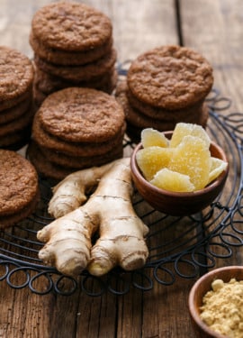 Stacks of Triple Ginger Snap Cookies on a wire rack, with a chunk of fresh ginger and bowls of crystalized and ground ginger on the side.