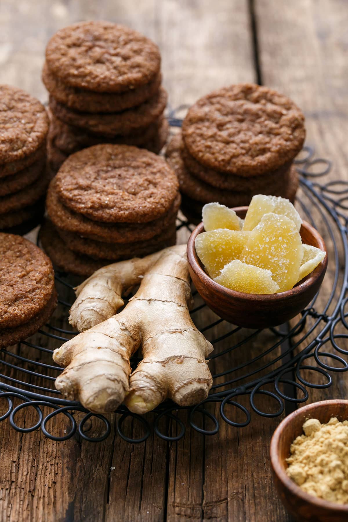 Stacks of Triple Ginger Snap Cookies on a wire rack, with a chunk of fresh ginger and bowls of crystalized and ground ginger on the side.