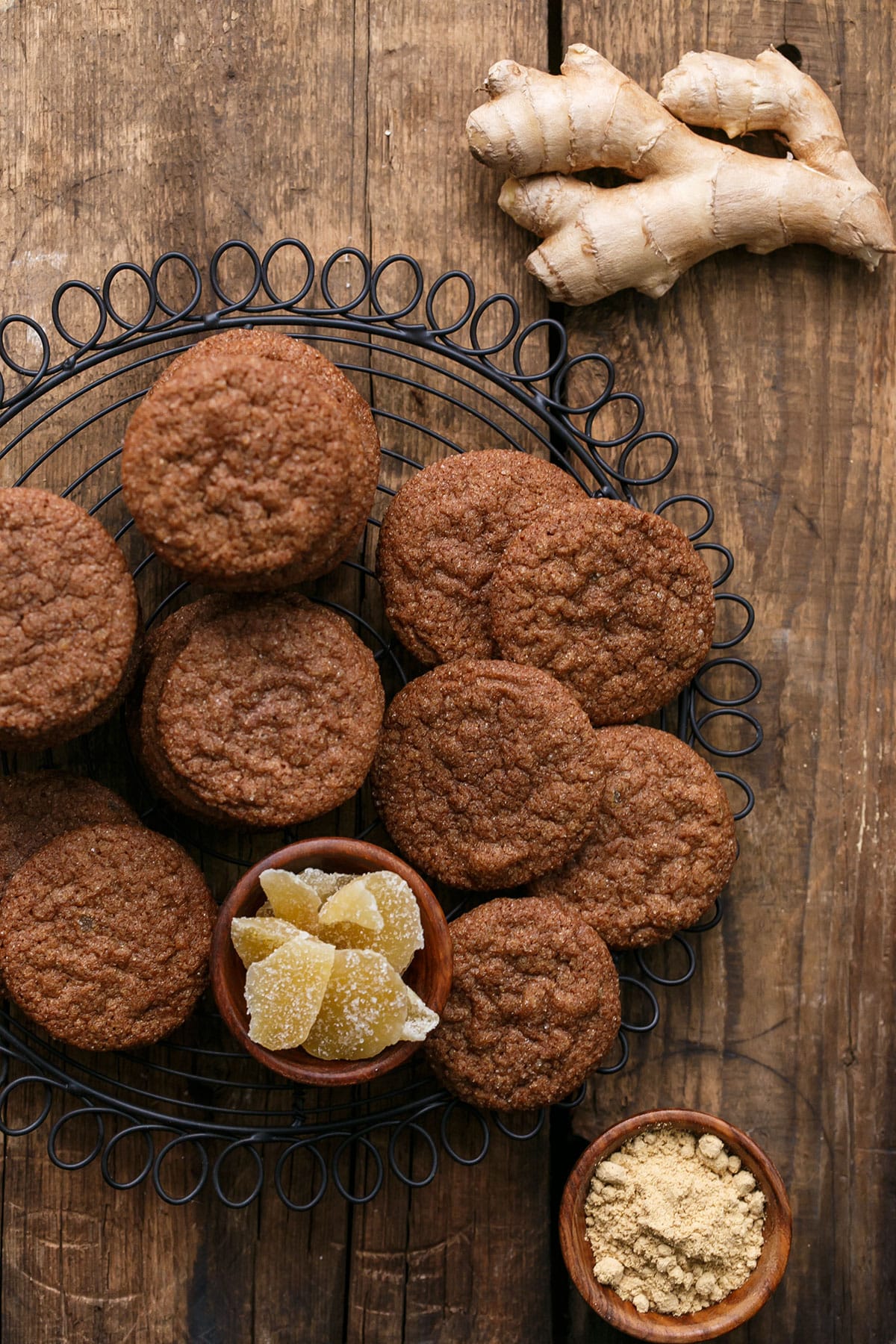 Overhead view of Triple Ginger Snap Cookies on a wire rack, with a wooden background and three kinds of ginger (fresh, ground, and crystalized).