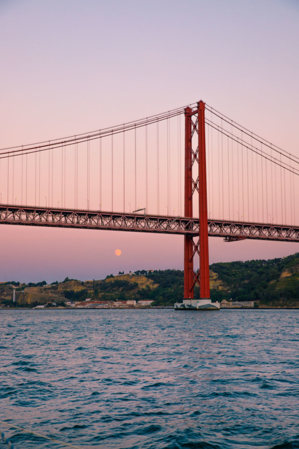 The moon visible at sunset underneath the 25 de Abril bridge in Lisbon, Portgual