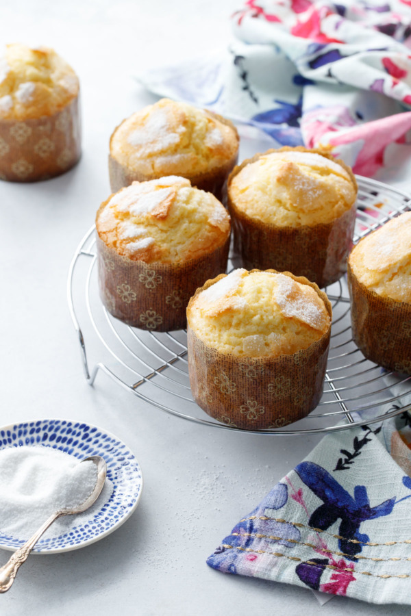 Five Portuguese rice muffins on a cooling rack with one in the background and a small dish of sugar 