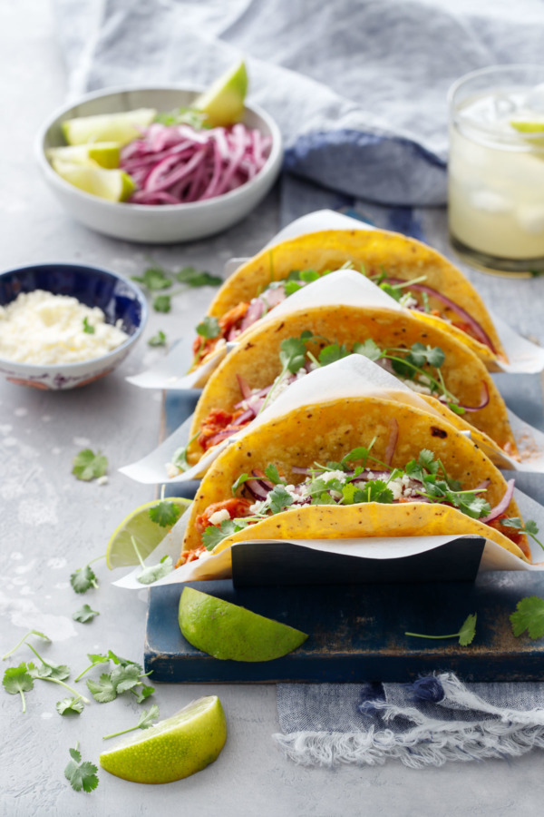 Three chicken tinga tacos in a stainless steel taco holder, with a bowl of crumbled cheese, pickled onions, and a margarita on the side.