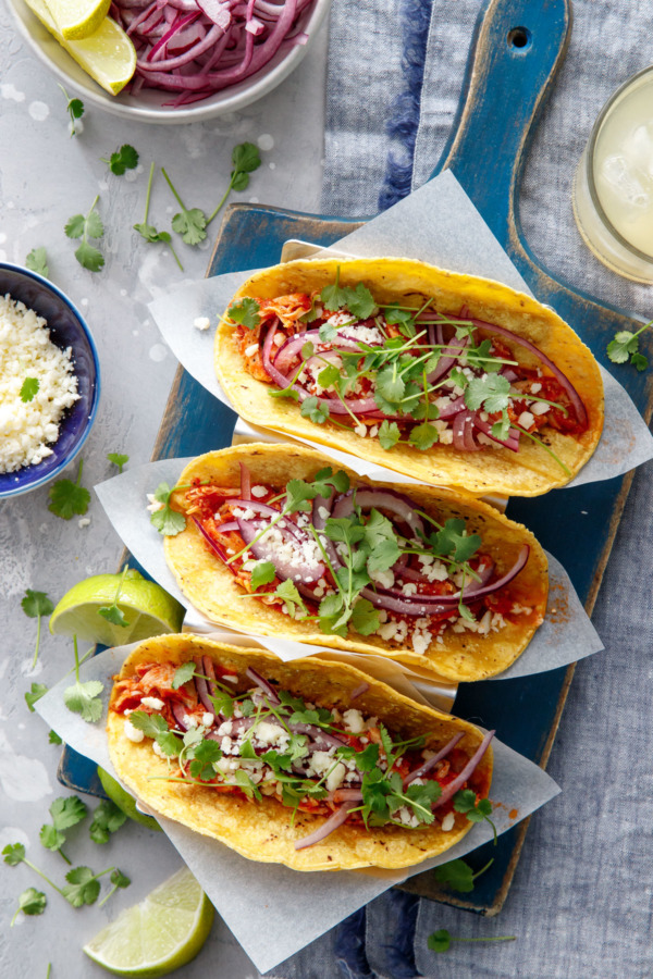 Overhead shot of Three chicken tinga tacos in a stainless steel taco holder, with a bowl of crumbled cheese, pickled onions, and a margarita on the side.