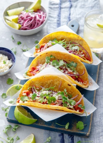 Three chicken tinga tacos in a stainless steel taco holder, with a bowl of crumbled cheese, pickled onions, and a margarita on the side.