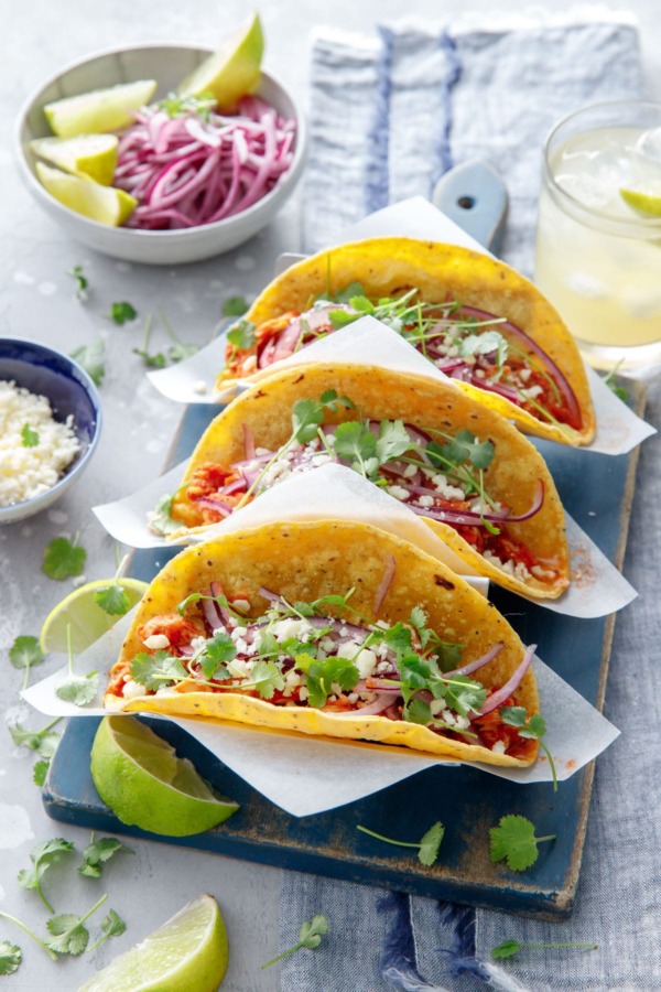 Three chicken tinga tacos in a stainless steel taco holder, with a bowl of crumbled cheese, pickled onions, and a margarita on the side.