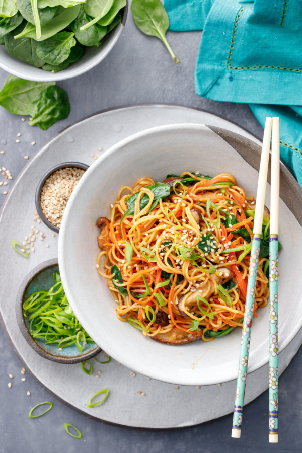 Overhead view of a bowl of stir fry noodles with chopsticks, and bowls of green onion, sesame seeds and fresh spinach on the side.