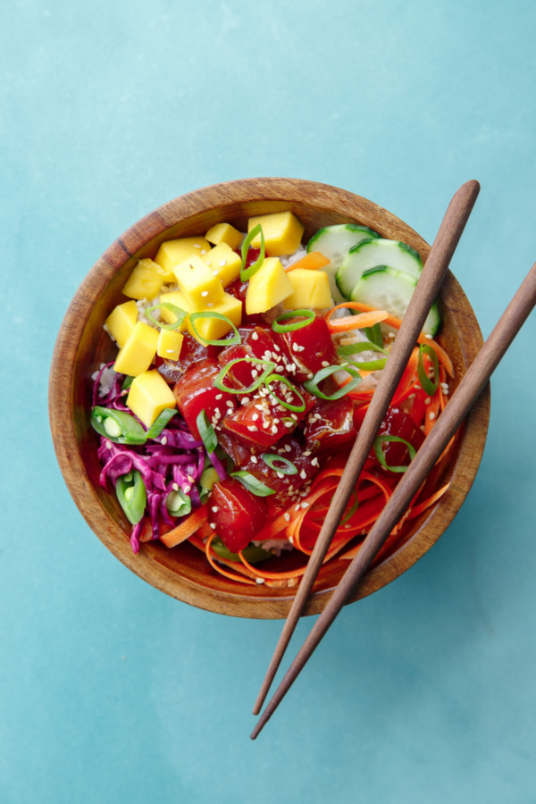 Overhead shot of a single wooden bowl with Ahi Mango Poke and chopsticks, on a turquoise background.